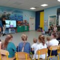 Zdjęcie przedstawia Panią Agnieszkę, która wskazuje na ekran, na którym, wyświetlana jest dziewczynka, która nakłada na swoją twarz kartkę, na której narysowana jest smutna mina. Dzieci siedzą na krzesełkach w półkole i z uwagą przyglądają się wyświetlanej grafice.