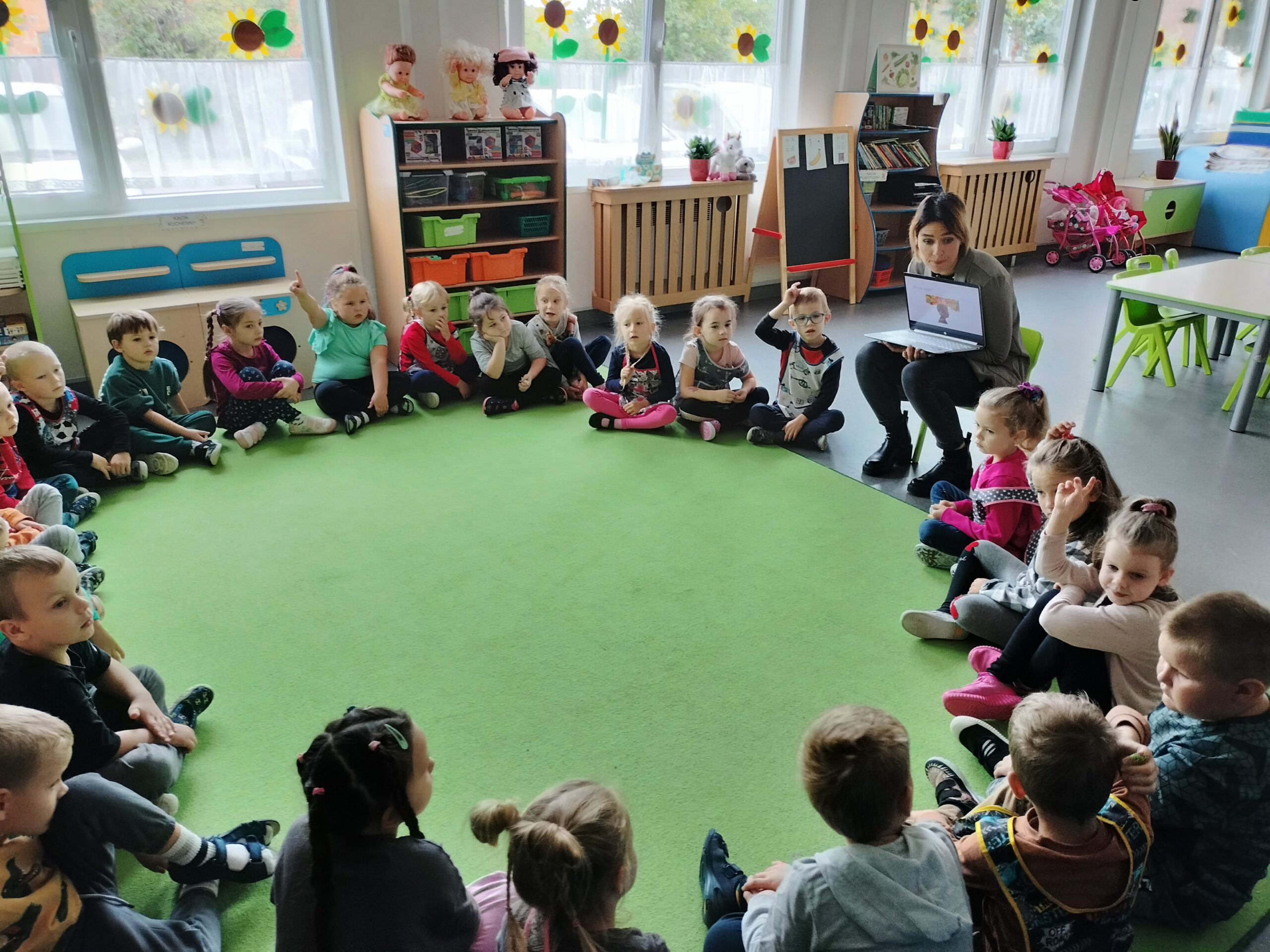 Zdjęcie przedstawia przedszkolaków, które siedzą po turecku tworząc koło. Wśród nich jest Pani Agnieszka, która naucza ich o wyrażaniu swoich emocji.
