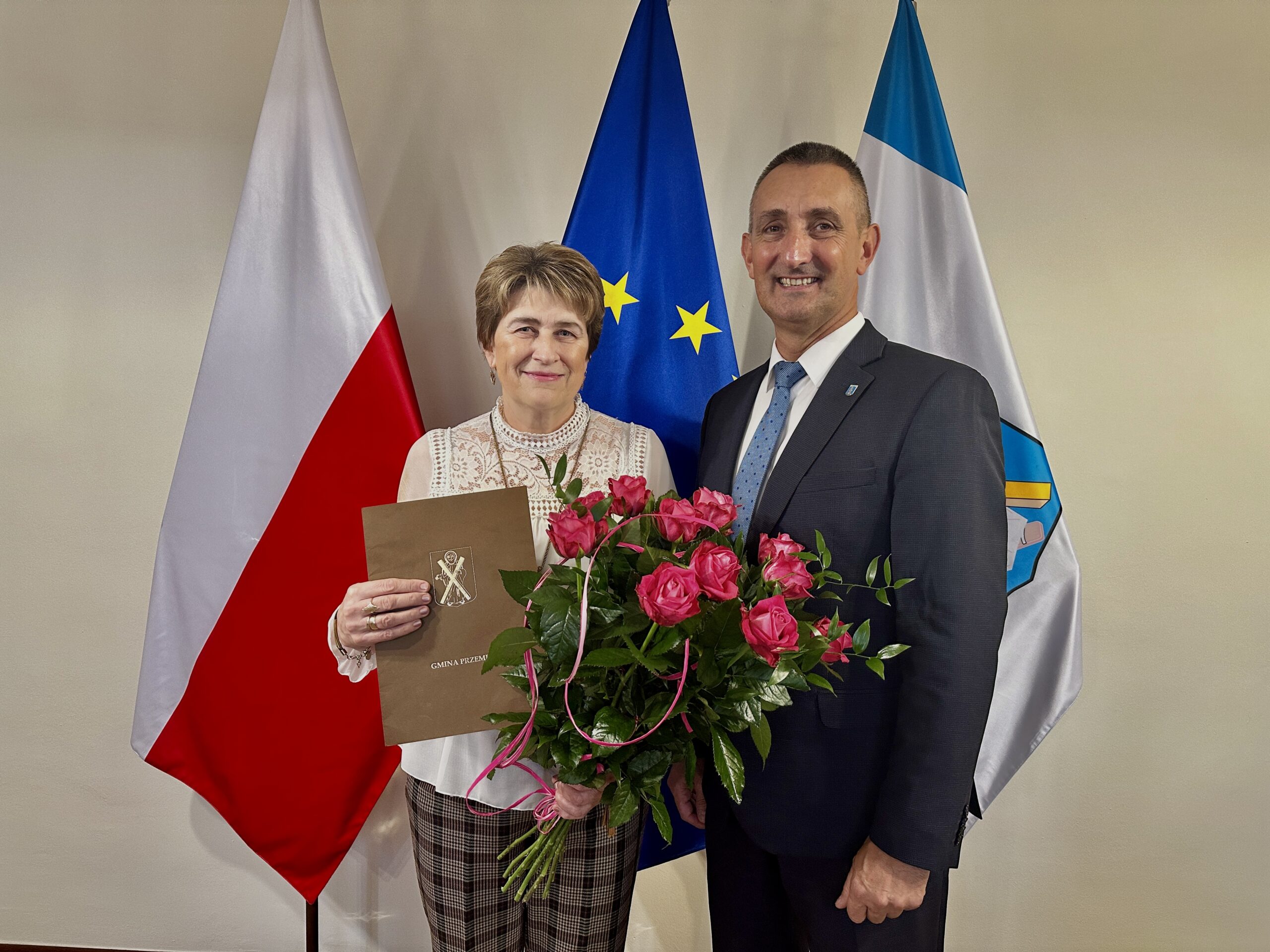 Na zdjęciu wójt Gminy Przemęt oraz Pani Jadwiga, która trzyma w ręce list gratulacyjny oraz bukiet różowych róż. Zdjęcie zrobiono w biurze wójta na tle flag.