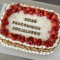 Na zdjęciu tort w jasnych kolorach, ozdobiony truskawkami oraz malinami. Na środku tortu widnieje napis: dzień pracownika socjalnego.