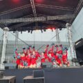 Zdjęcie przedstawia jedną z grup mażoretek Vena. Dziewczyny ubrane są w czerwone stroje ze srebrnymi dodatkami. Grupa występuje na scenie z pomponami.