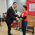 Zdjęcie przedstawia Wójta Gminy Przemęt, który wręcza paczkę wolontariuszce. W tle choinka ozdobiona papierowymi ozdobami, wokół której znajdują się paczki oraz materiałowa czerwona chusta z napisem szlachetna paczka.