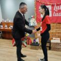 Zdjęcie przedstawia Wójta Gminy Przemęt, który wręcza paczkę wolontariuszce. W tle choinka ozdobiona papierowymi ozdobami, wokół której znajdują się paczki oraz materiałowa czerwona chusta z napisem szlachetna paczka.