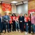 Zdjęcie przedstawia wszystkich zebranych pozujących do zdjęcia. W tle materiałowa, czerwona chusta z napisem szlachetna paczka.