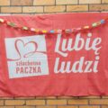 Zdjęcie przedstawia czerwoną chustę, którą zdobi kolorowy łańcuch wykonany z papieru. Na chuście widnieje napis "szlachetna paczka" oraz "Lubię ludzi".