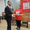 Zdjęcie przedstawia Wójta Gminy Przemęt rozmawiającego z wolontariuszką. W tle choinka ozdobiona papierowymi ozdobami, wokół której znajdują się paczki oraz materiałowa czerwona chusta z napisem szlachetna paczka.