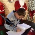 Zdjęcie przedstawia chłopaka, który długim niebieskim piórem podpisuje się w dzienniku św. Mikołaja. Na zdjęciu widać też Mikołaja.