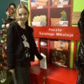 Zdjęcie przedstawia uśmiechniętą dziewczynkę, która wrzuca swój list do skrzynki z pocztą do św. Mikołaja. W kolejce czeka reszta dzieci na swoją kolej.