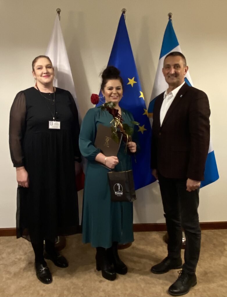 Zdjęcie przedstawia Wójta Gminy Przemęt, Kierownika Wydziału Oświaty, Promocji, Turystyki i Sportu Panią Martę Sobierajską oraz Panią Ewę Pelec, która trzyma w rękach czerwoną różę oraz akt nadania. Zdjęcie wykonane w biurze wójta.