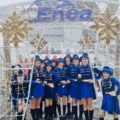 Zdjęcie przedstawia jedną z grup mażoretek Vena. Dziewczyny ubrane są w niebieskie stroje ze złotymi elementami. Pozują na tle ozdoby świątecznej.