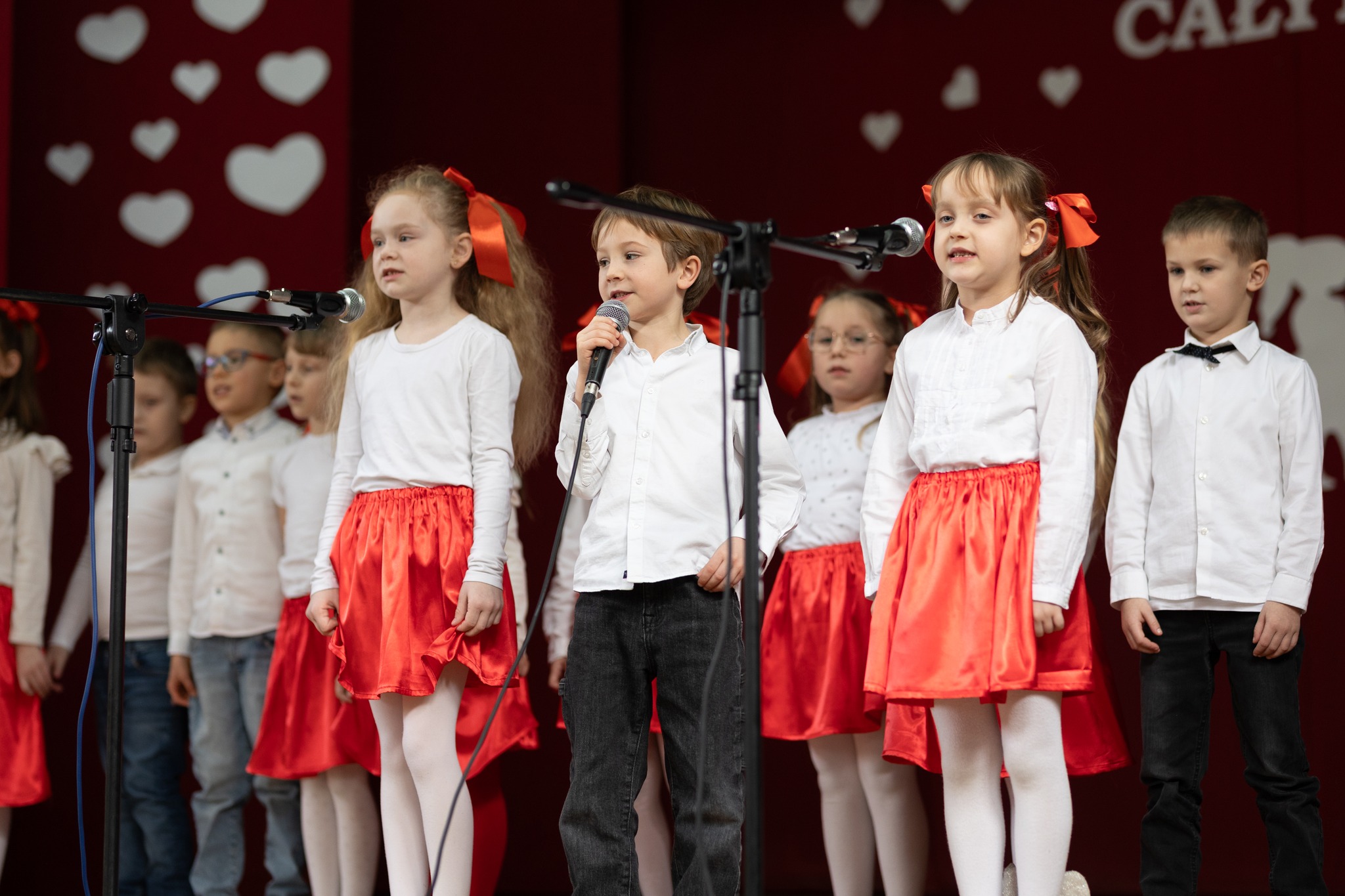 Zdjęcie przedstawia przedszkolaków śpiewających na scenie. Dziewczynki ubrane są w biało-czerwone stroje, a chłopcy w biało-czarne. Na głównym planie znajduje się chłopiec, który stoi pomiędzy dwiema dziewczynkami.