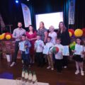 Wspólne zdjęcie laureatów z jury oraz organizatorami. Dzieci pozują z nagrodami. Wszyscy pozują na tle sceny, na której jest projektor, na którym widnieje napis: "XIII Gminny Konkurs Recytatorski Przedszkolaków". Scena ozdobiona jest kolorowymi dodatkami.