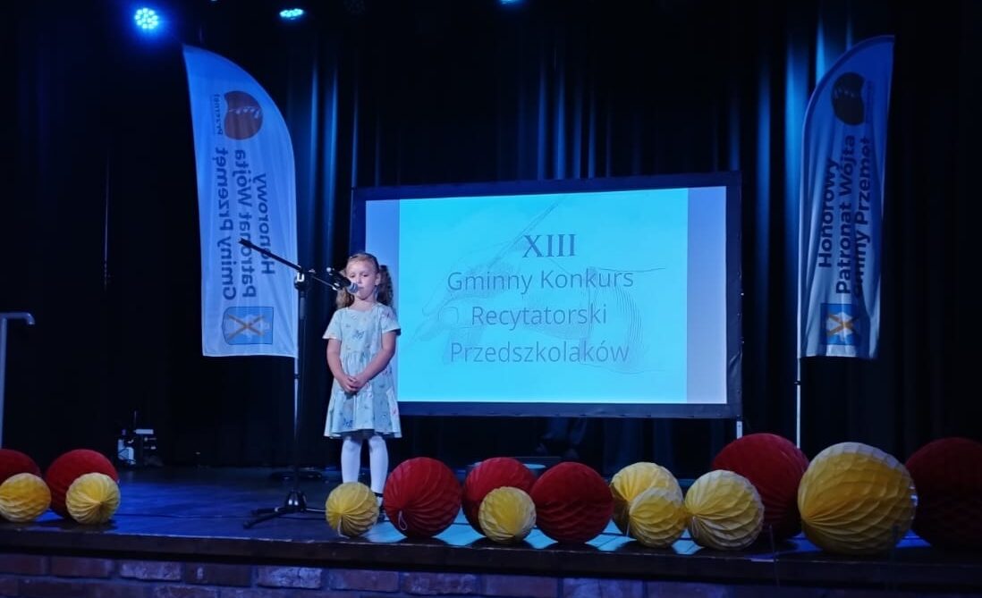 Na zdjęciu dziewczynka na scenie recytująca swój wiersz. Za nią projektor, na którym widnieje napis: "XIII Gminny Konkurs Recytatorski Przedszkolaków". Scena ozdobiona jest kolorowymi dodatkami. Na fotografii uchwycono także windery Honorowy Patronat Wójta Gminy Przemęt.