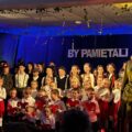 Zdjęcie przedstawia dzieci w wieku szkolnym i przedszkolnym. Wszyscy stoją razem na scenie. Przedszkolaki trzymają w rękach duże czerwone serduszka z papieru. Młodsi ubrani są w barwy Polski, natomiast starsi prezentują się w strojach charakterystycznych dla tamtych czasów oraz w mundurach.