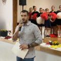 Mężczyzna przemawia przez mikrofon, w tle na scenie dzieci.