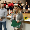 Kobieta przemawia przez mikrofon, w tle mężczyzna uśmiecha się. W tle dzieci na scenie.