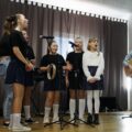 Dziewczynki stoją na scenie i śpiewają, z prawej mężczyzna gra na gitarze