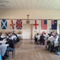 Na zdjęciu widać ścianę sali, na której wiszą flagi od prawej: Australii, Stanów Zjednoczonych, Anglii, Wielkiej Brytanii, Irlandii Północnej, Finlandii oraz Walii. Po bokach zdjęcia widać uczniów siedzących przy stołach i skupionych na teście.