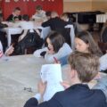 Na zdjęciu grupka uczniów pisząca test z języka angielskiego przy prostokątnych stołach. Sala została ozdobiona w papierowe flagi Wielkiej Brytanii i inne dodatki.