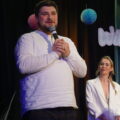 Mężczyzna przemawia przez mikrofon. W tle kobieta zerka na mężczyznę.