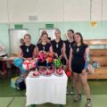 Na zdjęciu dziewczyny ze starszej klasy z szkoły w Mochach. Dziewczyny trzymają w rękach nagrody rzeczowe w postaci piłek do piłki nożnej. Przed nimi znajduje się stół, na którym są puchary oraz medale.