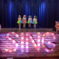 Zdjęcie przedstawia scenę, na przodzie której widnieje duży różowy napis w kropki: "SING", który jest podświetlony. Na scenie występuje czwórka dziewczynek, które ubrane są w wielobarwne spódniczki i białe bluzki. Każda z nich ma zrobione dwie kiteczki, które zostały ozdobione kolorowymi wstążkami.