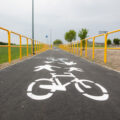 Zdjęcie przedstawia oznakowanie ścieżki rowerowej. Są to rowery oraz piesi. Wokół ścieżki rozciągają się żółte barierki.