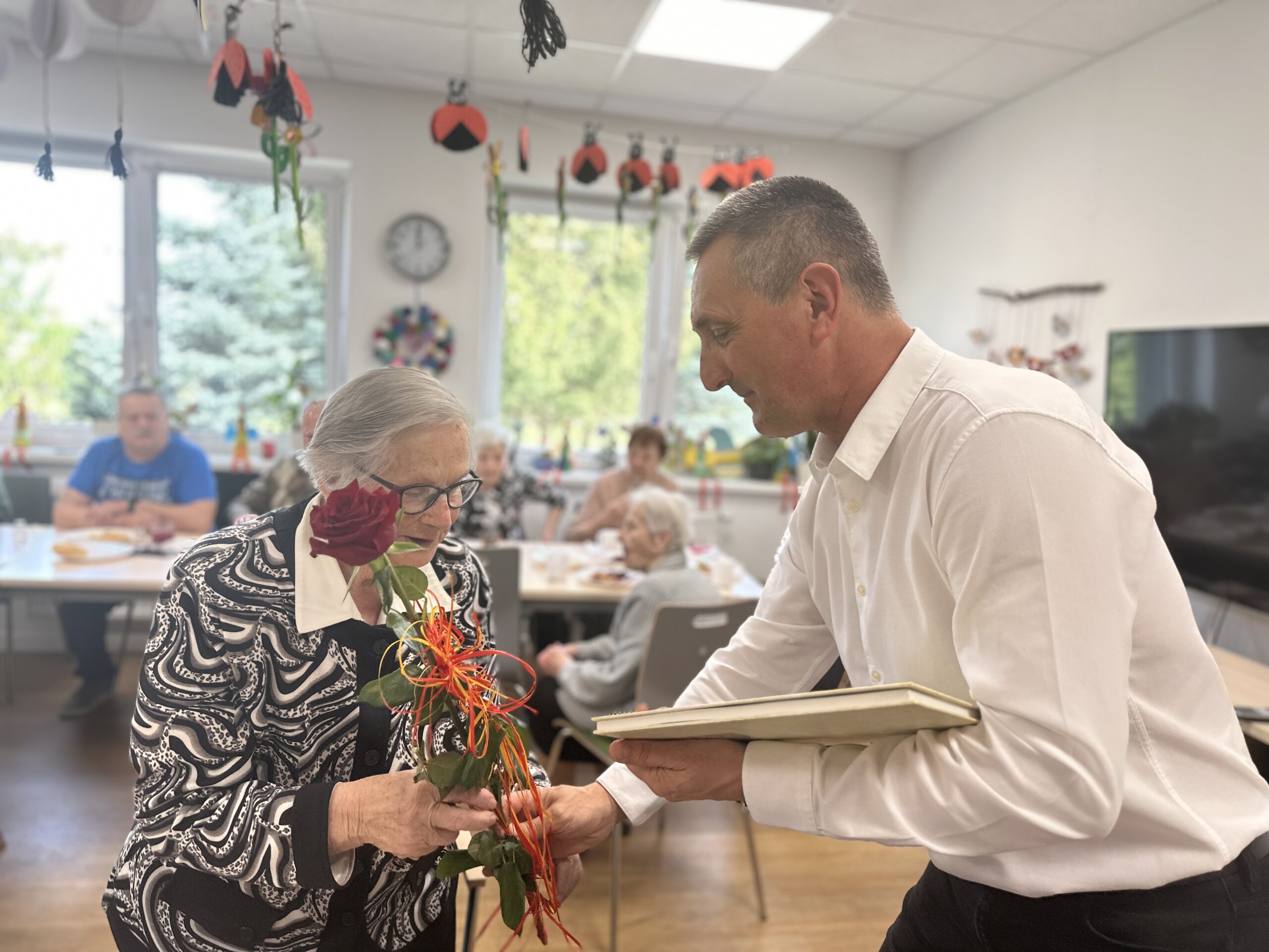 Na zdjęciu wójt Gminy Przemęt, który przekazuje Pani Irenie czerwoną różę oraz książkę. W tle pozostali seniorzy.