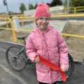 Zdjęcie przedstawia uśmiechniętą dziewczynkę ze szkoły w Radomierzu. Dziewczynka trzyma w ręce kawałek przeciętej wstęgi. W tle za nią znajduje się rower.