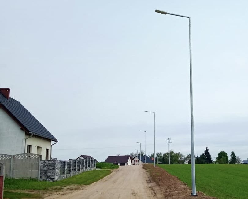 Zdjęcie przedstawia lampy ustawione wzdłuż ulicy Akacjowej w Radomierzu. Po drugiej stronie ulicy znajdują się domy. Zdjęcie wykonano w dzień.
