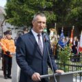 Wójt - mężczyzna przemawia przy mównicy, w tle sztandary oraz orkiestra