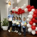 Na zdjęciu pozują przedszkolaki z Bucza z medalami, dyplomami oraz nagrodami wraz z przedszkolanką (opiekunem drużyny) na ściance ozdobionej balonami w barwach białych i czerwonych. Obok ścianki winder Honorowy Patronat Wójta Gminy Przemęt.