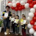 Na zdjęciu pozują przedszkolaki ze Starkowa z medalami, dyplomami oraz nagrodami wraz z przedszkolanką (opiekunem drużyny) na ściance ozdobionej balonami w barwach białych i czerwonych. Obok ścianki winder Honorowy Patronat Wójta Gminy Przemęt.