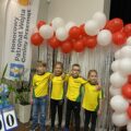 Na zdjęciu pozuje drużyna z przedszkola z Przemętu na tle ścianki. Dwie dziewczynki i dwóch chłopców. Dzieci ubrane są w jednakowe koszulki żółto- zielone, na których małym druczkiem widnieje czarny napis Przemęt. Na zdjęciu widać tez winder honorowy patronat Wójta Gminy Przemęt.