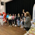Zdjęcie przedstawia Panią Elżbietę, która wygłasza przemowę powitalną. W ręce trzyma jeden z pucharów, który prezentuje wszystkim zebranym. Na zdjęciu znajduje się również reszta organizatorów oraz niektóre panie przedszkolanki.