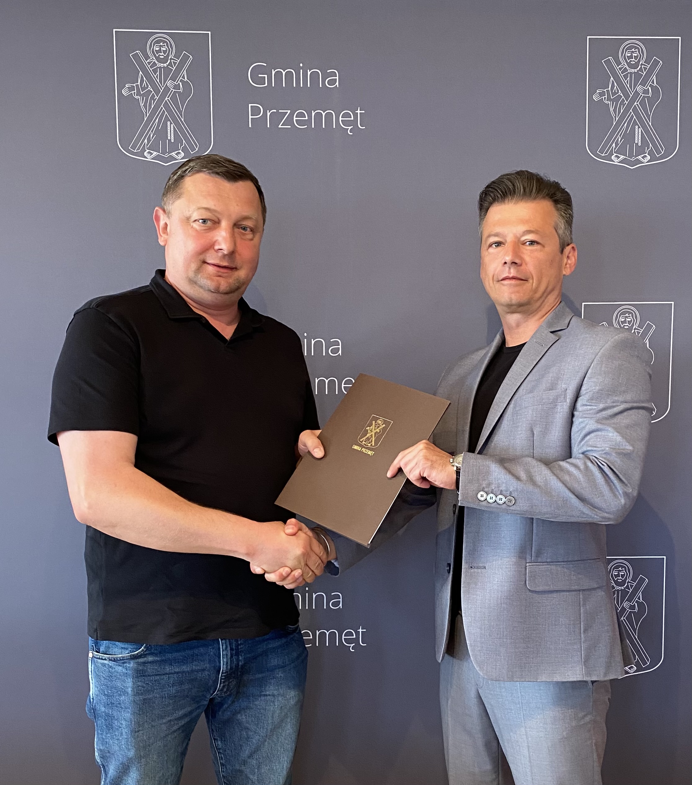 Na zdjęciu zastępca wójta Gminy Przemęt Waldemar Kalitka wraz z Sławomirem Białasikiem. Wyżej wymienieni podają sobie dłonie, a w rękach trzymają podpisaną umowę w teczce Gminy Przemęt. W tle ścianka z napisem Gmina Przemęt i herbem gminy, który przedstawia postać św. Andrzeja.