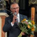 Wójt Frąckowiak przemawia przez mikrofon, w ręce trzyma kwiaty.