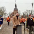 Konferansjer otwiera wydarzenie, w tle kościół. mażoretki, orkiestra i harcerze.