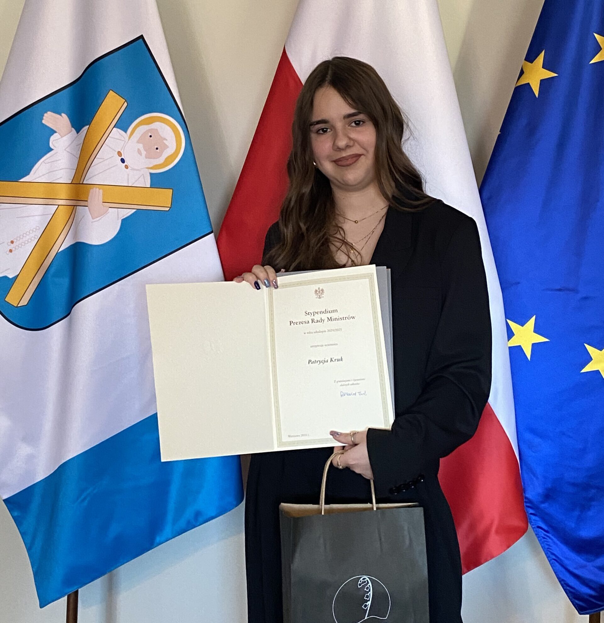 Na zdjęciu Patrycja Kruk pozuje wraz z listem gratulacyjnym - Stypendium Prezesa Rady Ministrów oraz upominkiem od wójta gminy Przemęt. W tle flagi: polska, europejska oraz z herbem gminy Przemęt.