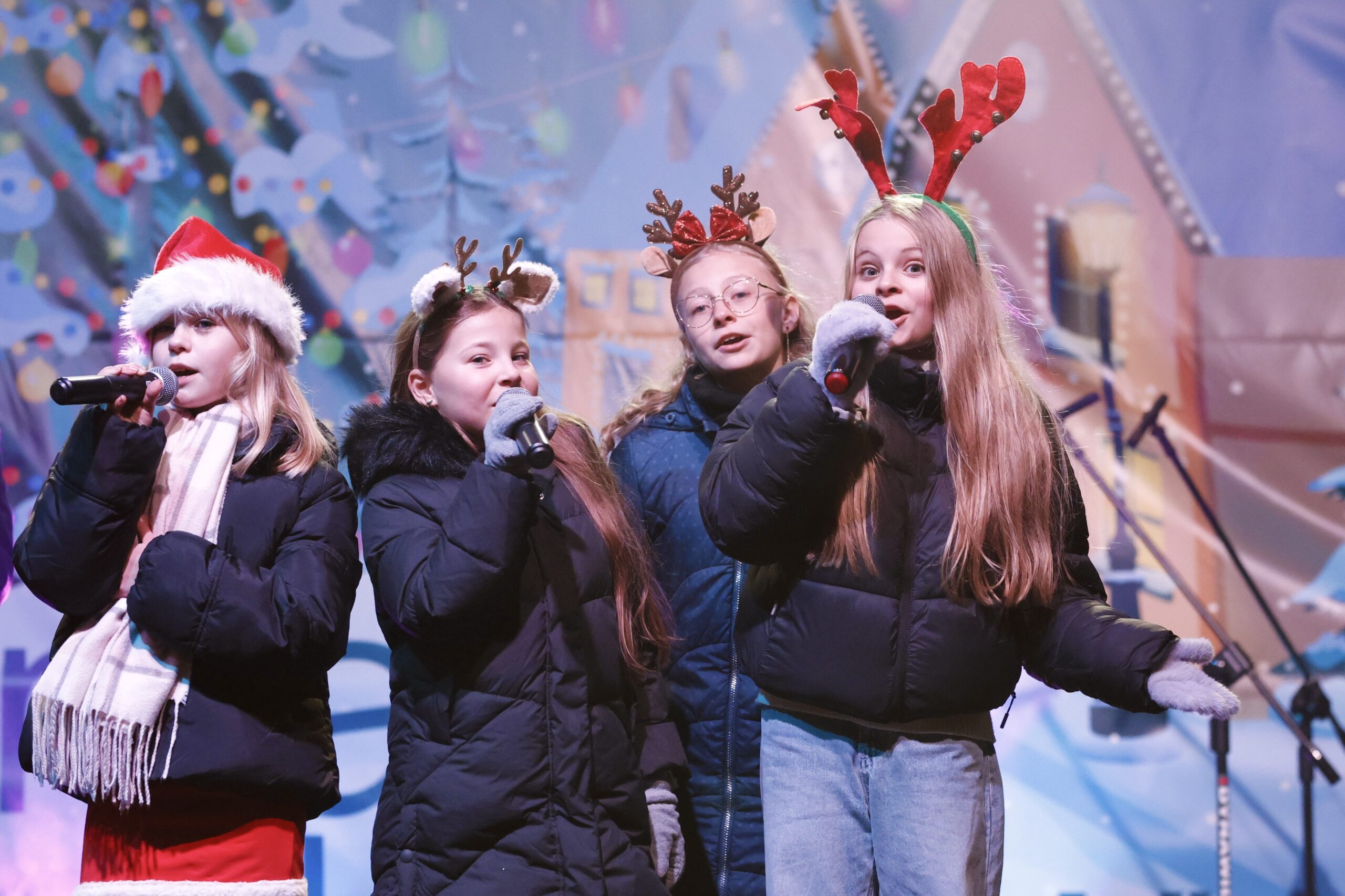 Dziewczynki w świątecznych strojach śpiewają przez mikrofony na scenie.