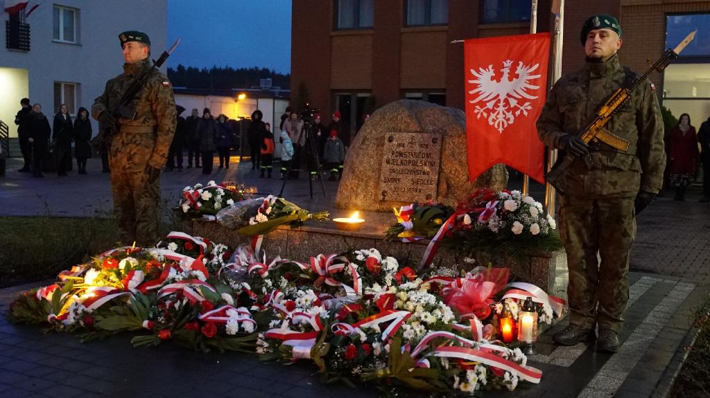 Na zdjęciu dwójka żołnierzy. Żołnierze stoją po obu stronach pomnika, wokół którego ułożono wiązanki i znicze. Na fotografii widoczna także flaga powstania wielkopolskiego.