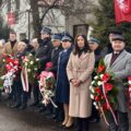 Na zdjęciu osoby uczestniczące na uroczystości na rynku. Niektóre z nich trzymają wiązanki kwiatów.