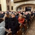 Zdjęcie przedstawia osoby siedzące w ławkach podczas mszy świętej.