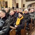 Zdjęcie przedstawia osoby siedzące w ławkach podczas mszy świętej.