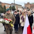 Na zdjęciu żołnierz składa, odebraną od delegacji, wiązankę kwiatów pod pomnikiem na rynku. W tle delegacje.
