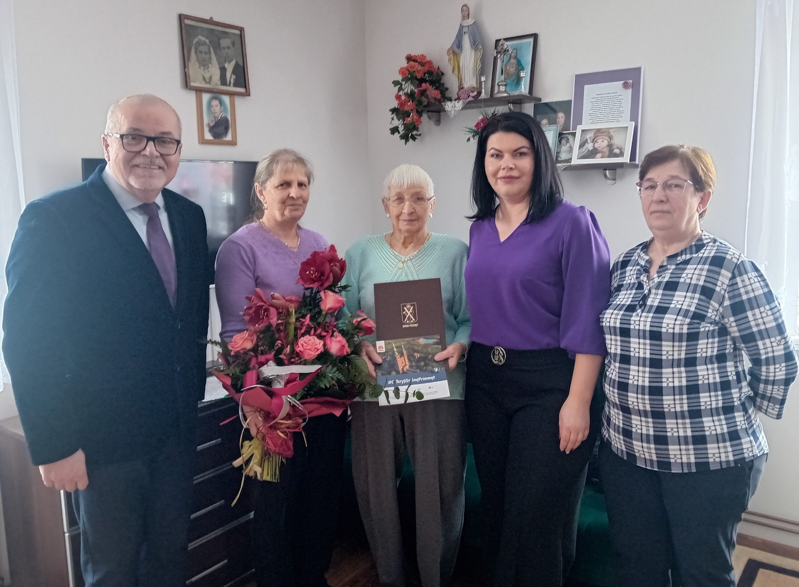 Zdjęcie przedstawia jubilatkę z córkami, przewodniczącą rady gminy Przemęt Moniką Maćkowiak wraz z Kierownikiem Urzędu Stanu Cywilnego Leszkiem Anglartem. Jubilatka pozuje z listem gratulacyjnym i książeczką o gminie Przemęt, a jedna z jej córek z bukietem kwiatów.