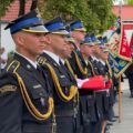 Zdjęcie przedstawia strażaków ustawionych w rzędzie. Zdjęcie wykonano z profilu.