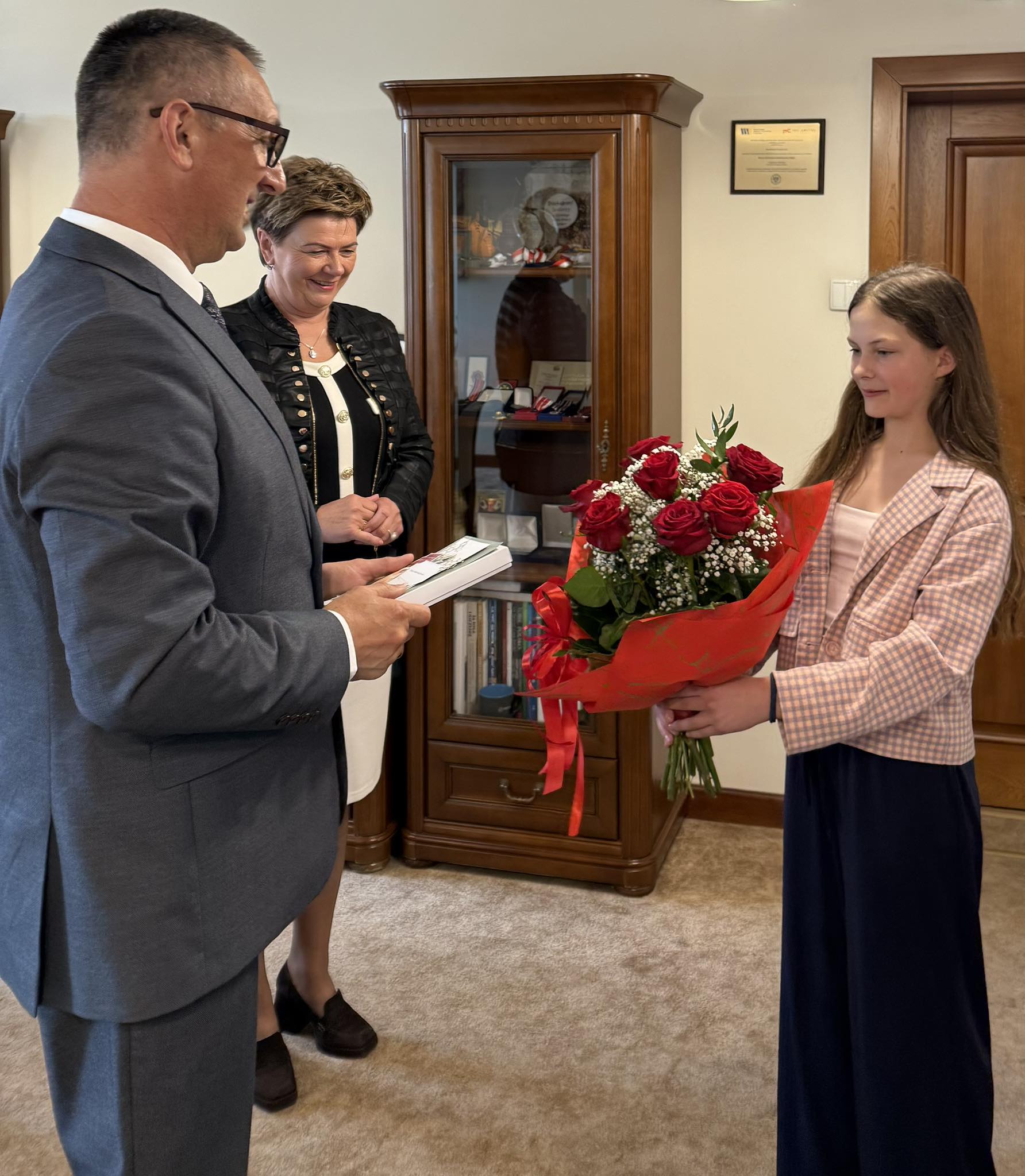 Na fotografii wójt gminy Janusz Frąckowiak, dyrektor szkoły Małgorzata Zielińska oraz uczennica szkoły. Wójt trzyma w ręce upominek. Uczennica wręcza wójtowi bukiet czerwonych róż.