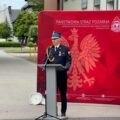 Na zdjęciu komendant powiatowy państwowej straży pożarnej Damian Glapiak wygłasza przemowę. W tle czerwona ścianka z orłem oraz napisem: "Państwowa Straż Pożarna. Komenda Powiatowa Państwowej Straży Pożarnej w Wolsztynie".