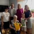 Wspólne zdjęcie, na którym znajduje się wójt, zastępca kierownika urzędu stanu cywilnego pani Irena oraz jubilatka wraz z córką oraz wnuczką i prawnukiem. Jubilatka trzyma bukiet kwiatów i list.